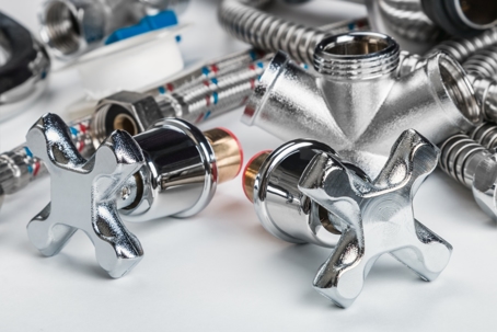 Close-up of shiny chrome plumbing fittings, valves, and flexible hoses arranged on a white surface, representing professional plumbing tools and supplies.